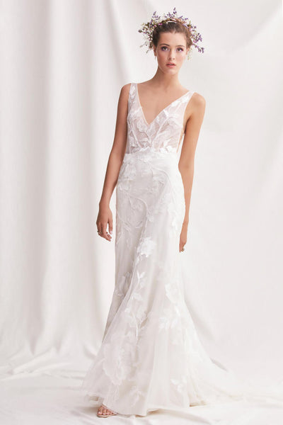 A woman in an elegant white Willowby by Watter Honor 52122 - Sample Size fit and flare gown with floral motifs stands against a plain background, adorned with a flower crown from Bergamot Bridal.