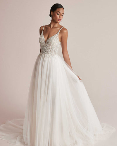 A woman in an elegant white Justin Alexander Cady Dress - Off The Rack with detailed embroidery on the bodice and a flowing English net skirt, standing against a soft pink background from Bergamot Bridal.