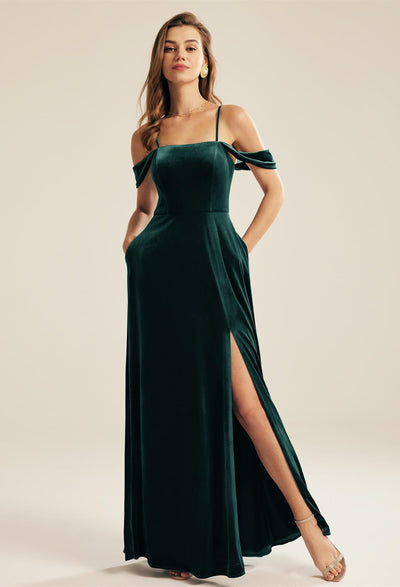 Woman in an elegant dark green Lowell velvet bridesmaid dress with off-shoulder sleeves and a high slit, standing confidently against a neutral background, purchased from Bergamot Bridal, one of the premier bridal shops in London.