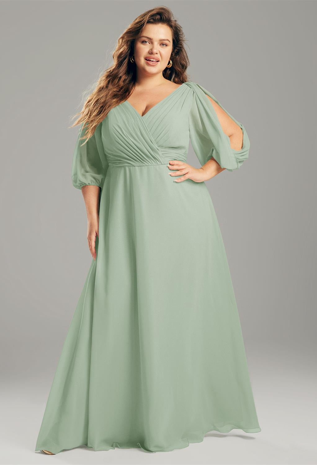 A woman in an elegant sage green Polly - Chiffon Bridesmaid Dress from Bergamot Bridal poses confidently against a gray background.