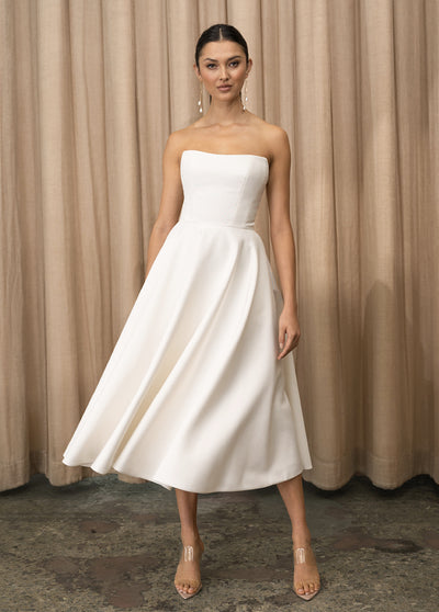 A model in a white strapless dress, possibly a Carrie - Jenny Yoo Little White Dress by Bergamot Bridal, posing in front of a curtain.