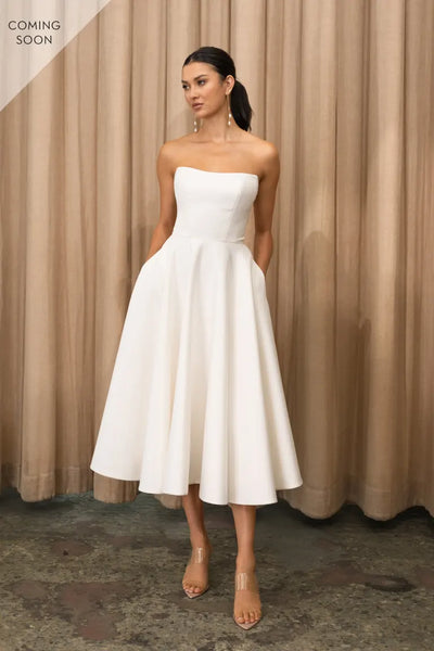 A woman in a strapless white midi dress from Bergamot Bridal stands against a draped beige background, wearing clear heeled sandals.