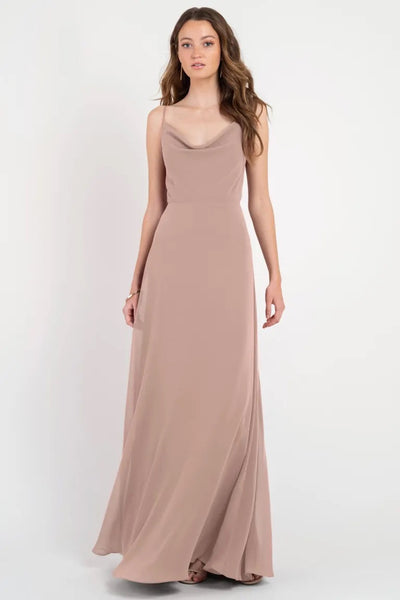 A woman in a long, flowing beige Colby - Jenny Yoo Bridesmaid Dress with a cowl neckline stands against a plain background from Bergamot Bridal.