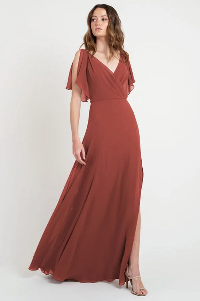 A woman in a beautiful, rust-colored evening gown with flutter sleeves and a v-neckline stands against a plain background - Hayes - Bridesmaid Dress by Jenny Yoo from Bergamot Bridal.