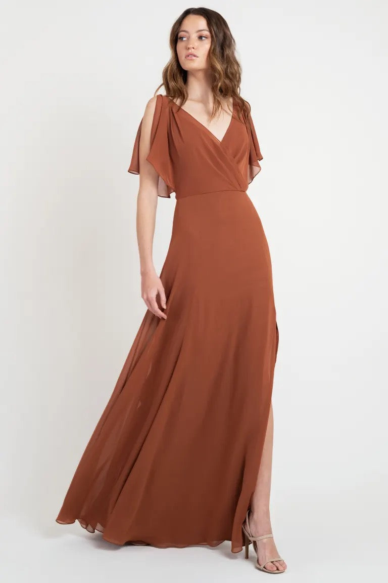 Woman in a beautiful rust-colored evening gown with flutter sleeves posing against a neutral backdrop, wearing the Hayes Bridesmaid Dress by Jenny Yoo from Bergamot Bridal.