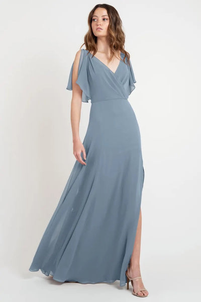 A woman models a beautiful floor-length blue Hayes - Bridesmaid Dress by Jenny Yoo with a slit and flutter sleeves from Bergamot Bridal.