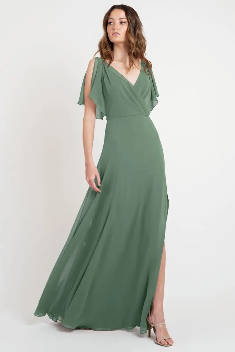 Woman posing in an elegant green evening dress with flutter sleeves and a high slit, the Hayes Bridesmaid Dress by Jenny Yoo from Bergamot Bridal.