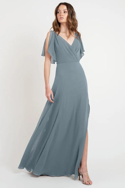 Woman modeling a Hayes - Bridesmaid Dress by Jenny Yoo, an elegant grey evening gown with flutter sleeves and a slit from Bergamot Bridal.