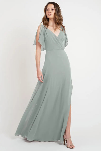 Woman in a beautiful, elegant Hayes - Bridesmaid Dress by Jenny Yoo in light green with flutter sleeves and a slit, posing for a Bergamot Bridal fashion display.
