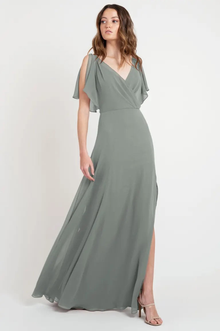Woman posing in a beautiful sage green maxi dress with a v-neckline and flutter sleeves, sample size 24 from Bergamot Bridal's Hayes - Bridesmaid Dress by Jenny Yoo.