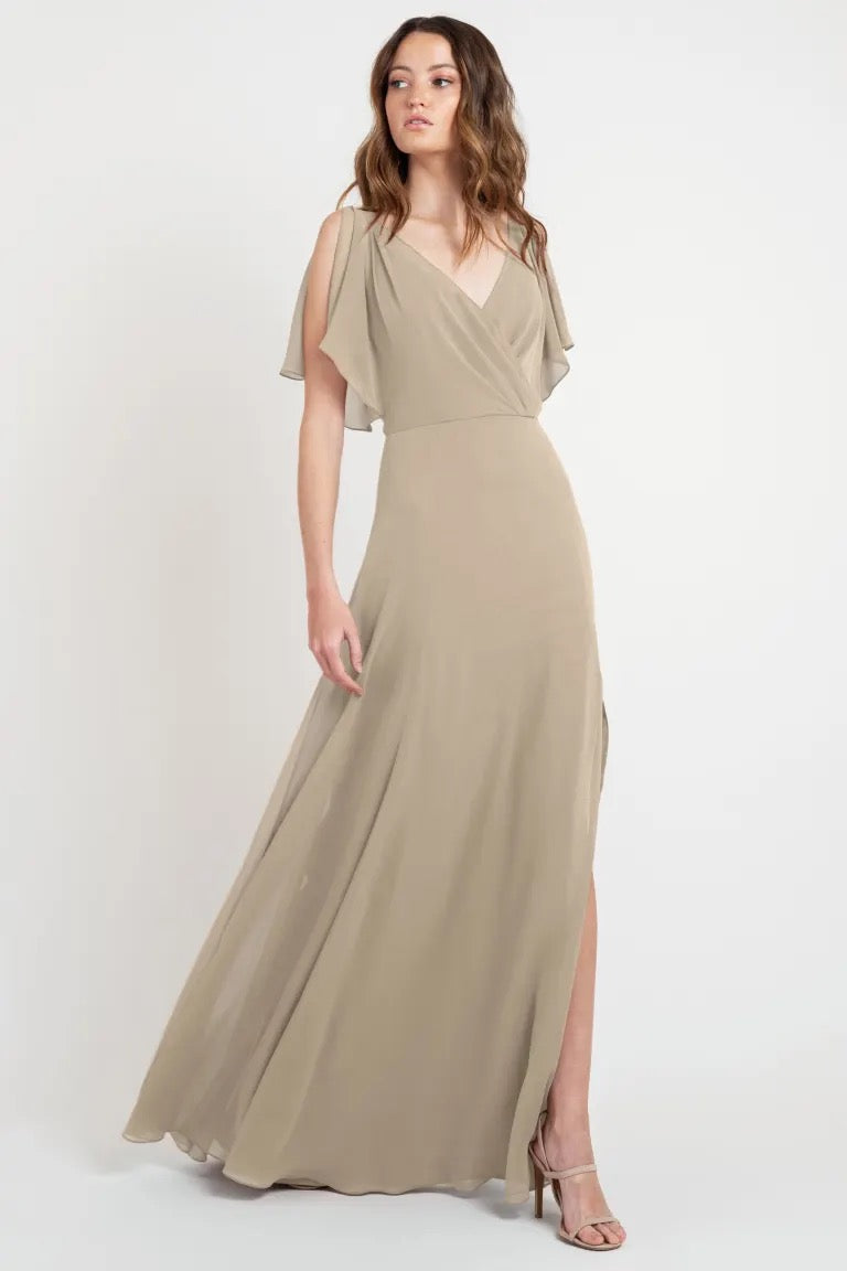 Woman posing in a flowing beige goddess dress with a v-neckline and flutter sleeves, the Hayes bridesmaid dress by Jenny Yoo at Bergamot Bridal.