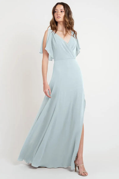 A woman in a beautiful light blue Hayes bridesmaid dress by Jenny Yoo with flutter sleeves and a slit, standing against a neutral background from Bergamot Bridal.