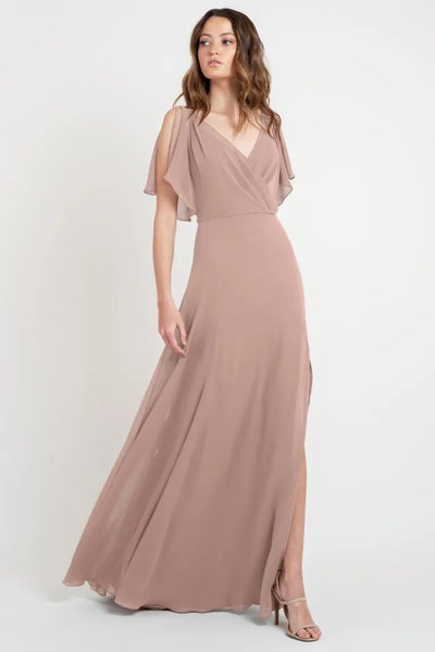 A woman in a beautiful dress, an elegant, flowing, dusky pink Hayes - Bridesmaid Dress by Jenny Yoo gown with flutter sleeves and v-neckline, standing against a plain background from Bergamot Bridal.