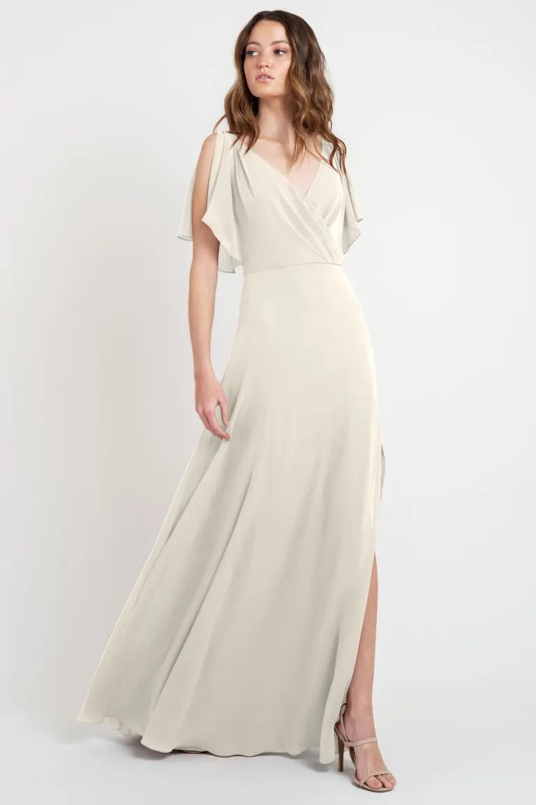 A woman posing in a Hayes - Bridesmaid Dress by Jenny Yoo with flutter sleeves and a flowing cream-colored evening gown with a slit, store sample size 24 from Bergamot Bridal.