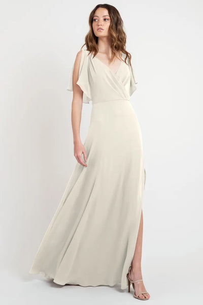 A woman posing in a Hayes - Bridesmaid Dress by Jenny Yoo with flutter sleeves and a flowing cream-colored evening gown with a slit, store sample size 24 from Bergamot Bridal.