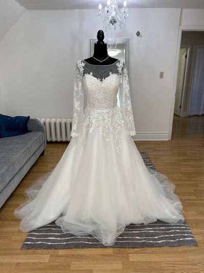 A Bridalane A-line Lace Long Sleeve Dress - Off The Rack displayed on a mannequin in a bright room with a chandelier and grey couch in the background.