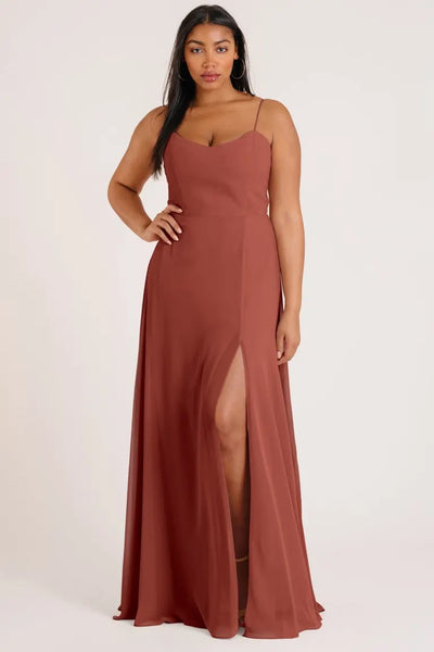 A woman in an elegant rust-colored luxe chiffon Kiara bridesmaid dress by Jenny Yoo with a slit on the side posing for the camera from Bergamot Bridal.