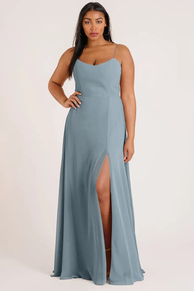 Woman posing in a luxe chiffon, blue Kiara - Bridesmaid Dress by Jenny Yoo with a scoop neck and a high slit from Bergamot Bridal.