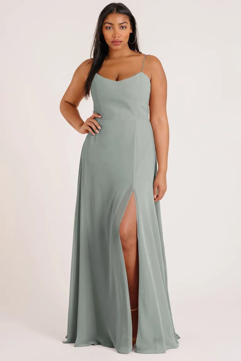 A woman modeling a sage green Kiara Bridesmaid Dress by Jenny Yoo with a luxe chiffon A-line skirt and a thigh-high slit from Bergamot Bridal.