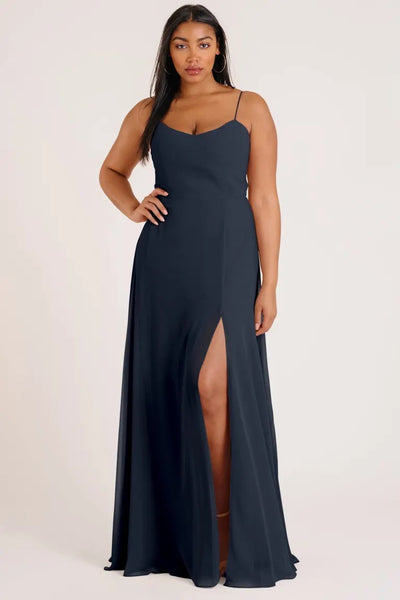 A woman posing in a Jenny Yoo Bridesmaid Dress by Kiara, an A-line skirt with a luxe chiffon navy blue evening gown featuring a thigh-high slit from Bergamot Bridal.