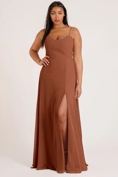 A woman in a luxe chiffon Kiara - Bridesmaid Dress by Jenny Yoo with an A-line skirt and a slit on the side standing against a light background from Bergamot Bridal.