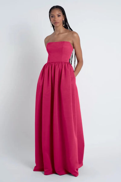 A woman in a strapless Laney bridesmaid dress by Jenny Yoo made of Luxe Faille fabric stands against a plain white background.