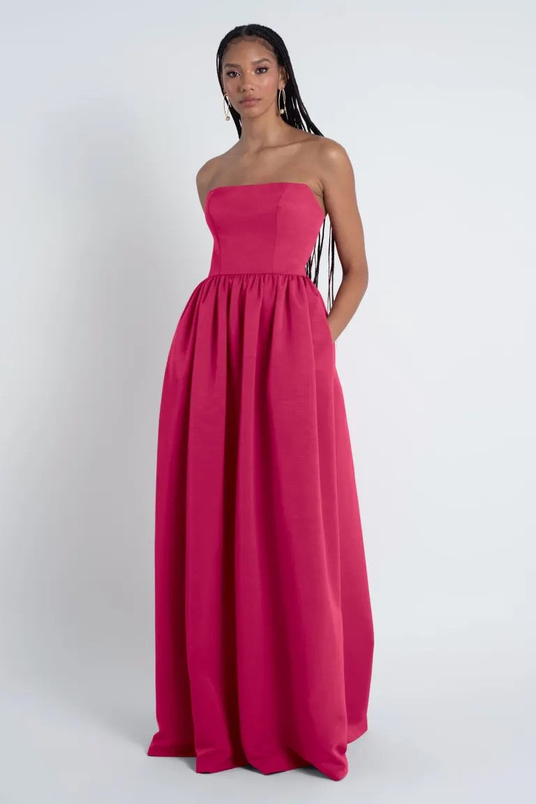 A young woman stands in a studio, wearing an elegant strapless pink Jenny Yoo Laney Bridesmaid Dress with a gathered skirt, looking directly at the camera from Bergamot Bridal.