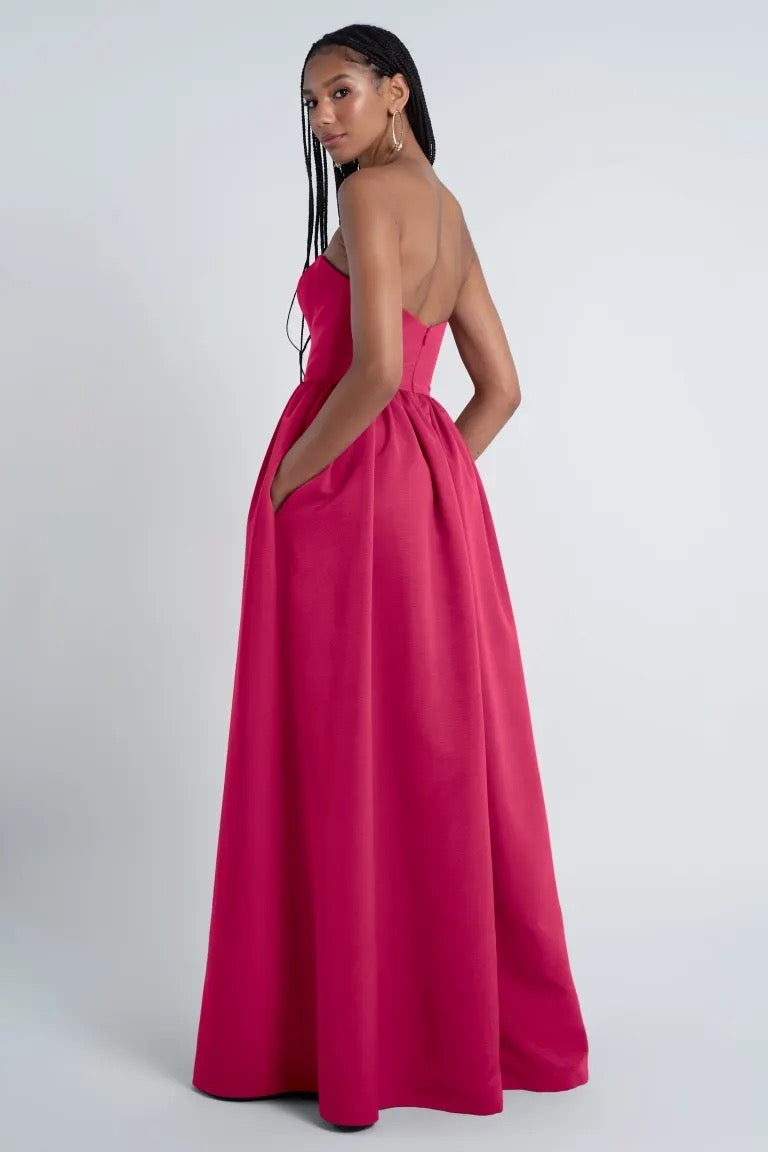 A woman in a vibrant pink Bridesmaid Dress by Jenny Yoo with pockets stands with her back turned, looking over her shoulder.