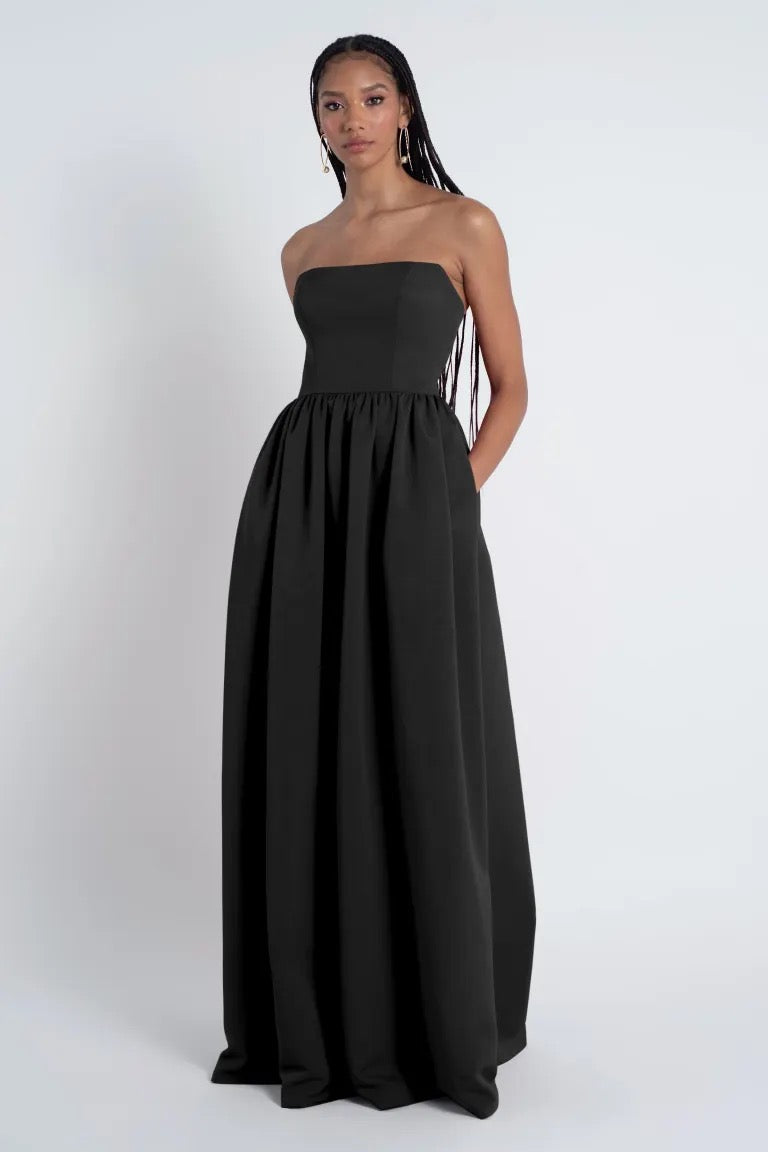 A woman in a strapless black Laney - Bridesmaid Dress by Jenny Yoo made of Luxe Faille fabric stands against a plain background, her hair styled in a long braid.