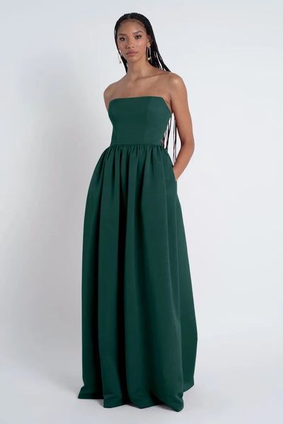 A woman in a strapless dark green Laney - Bridesmaid Dress by Jenny Yoo dress with a straight neckline stands against a gray background, her hair styled in long braids.