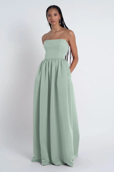 A woman in a Laney - Bridesmaid Dress by Jenny Yoo from Bergamot Bridal, in light green strapless gown with a gathered skirt, stands against a white background, looking directly at the camera.