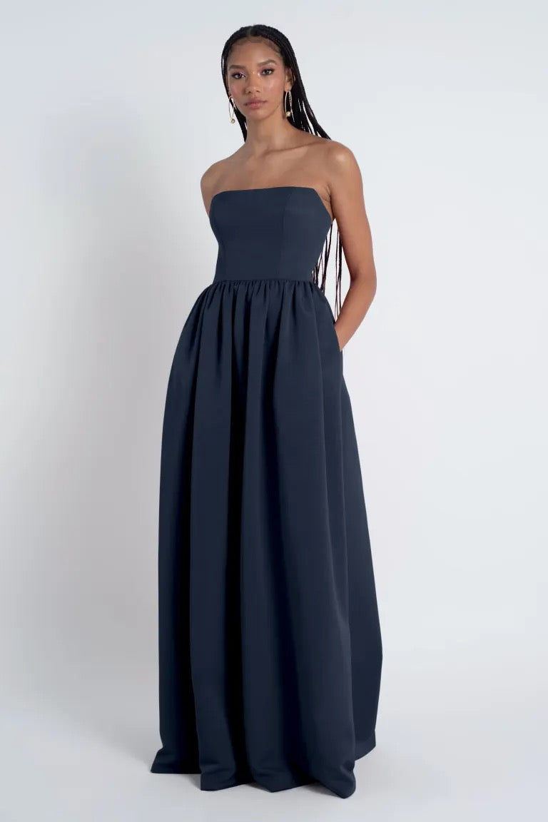 An elegant black woman posing in a navy Laney Bridesmaid Dress by Jenny Yoo with a straight neckline and strapless maxi length, featuring long braided hair, against a plain gray background.