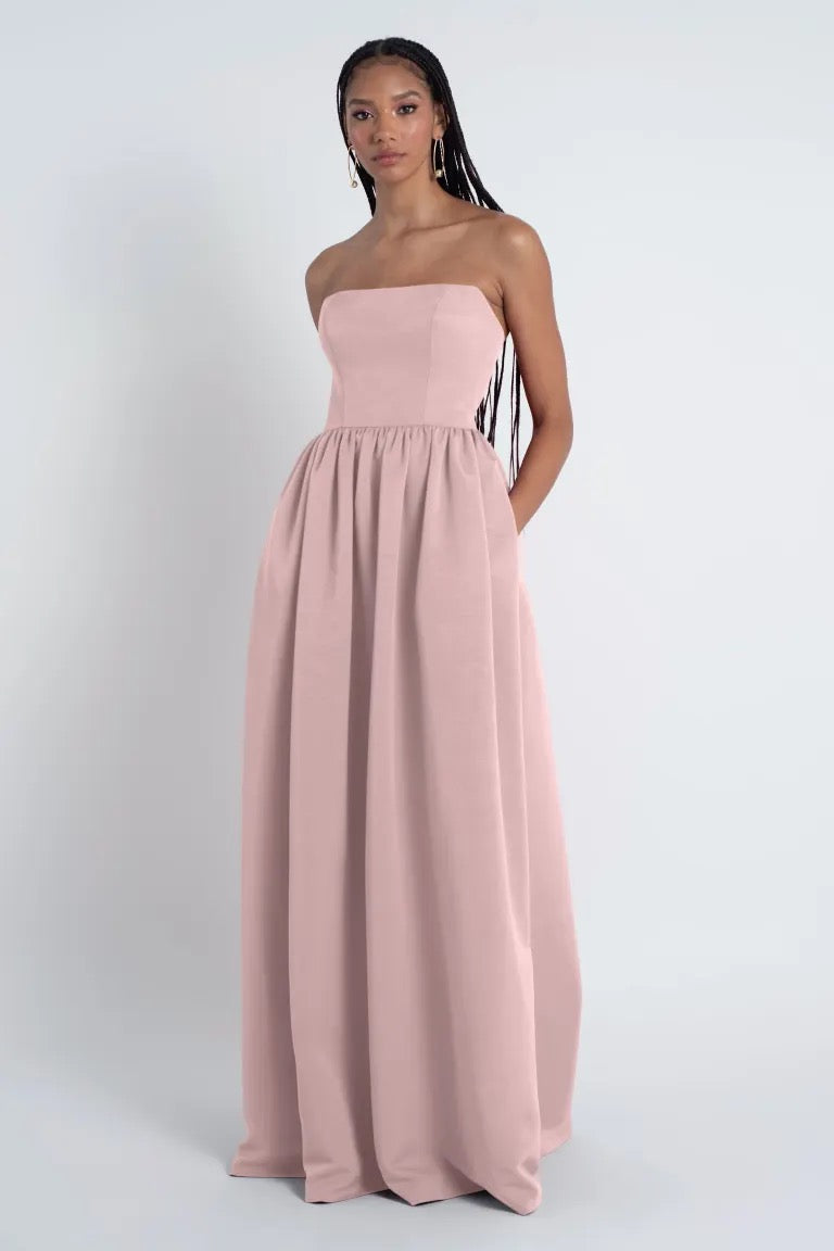 A young woman wearing a pale pink strapless Bridesmaid Dress by Jenny Yoo with a straight neckline, standing against a plain white background from Bergamot Bridal.