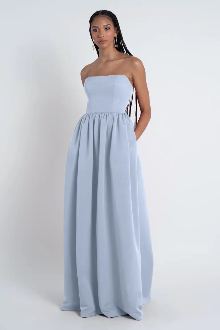 A young woman stands against a plain background, wearing a light blue Laney - Bridesmaid Dress by Jenny Yoo with a gathered skirt, looking directly at the camera.