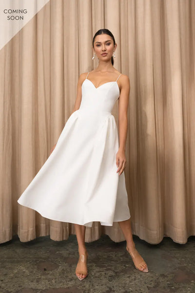 A woman wearing a white Luella - Jenny Yoo Little White Dress from Bergamot Bridal is standing in front of a curtain.