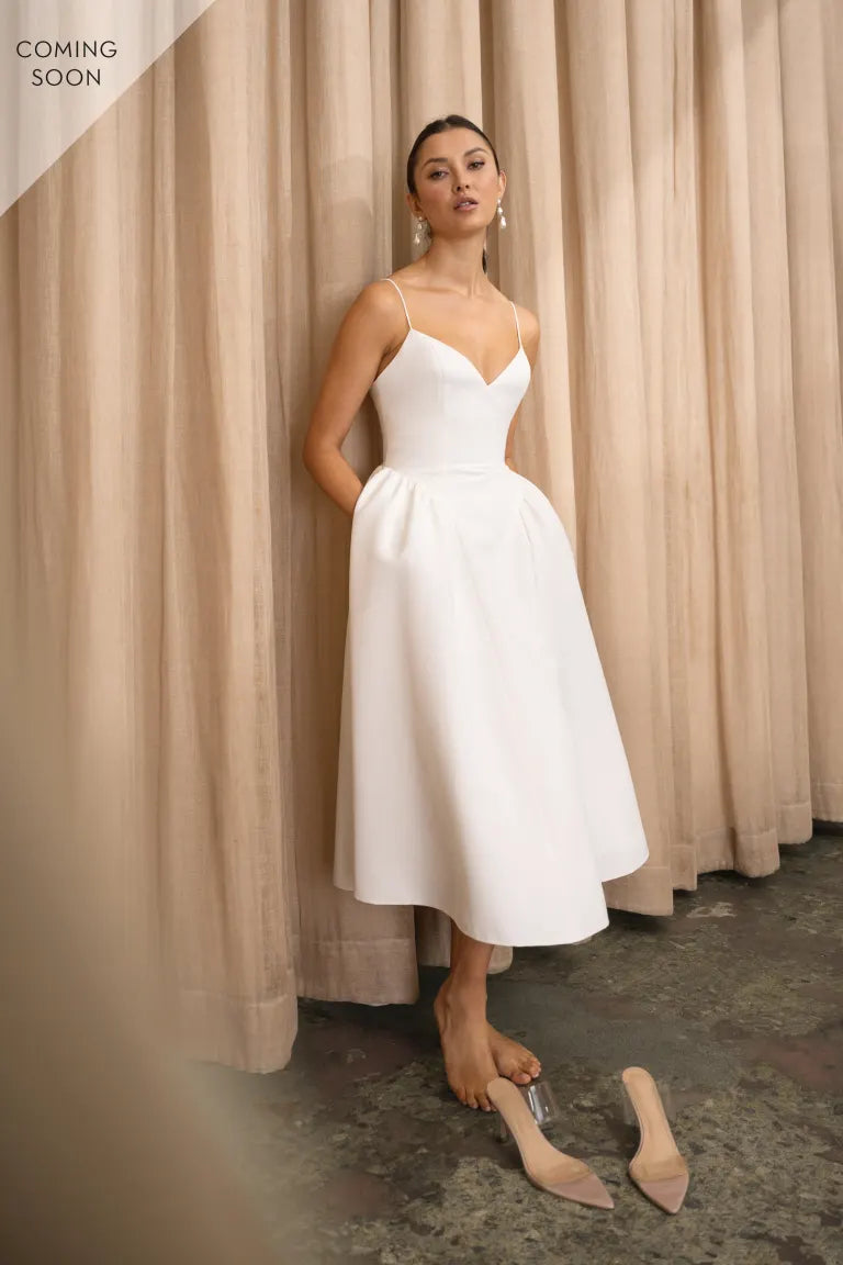 A woman in a wedding dress, the Luella - Jenny Yoo Little White Dress by Bergamot Bridal, standing next to a curtain.
