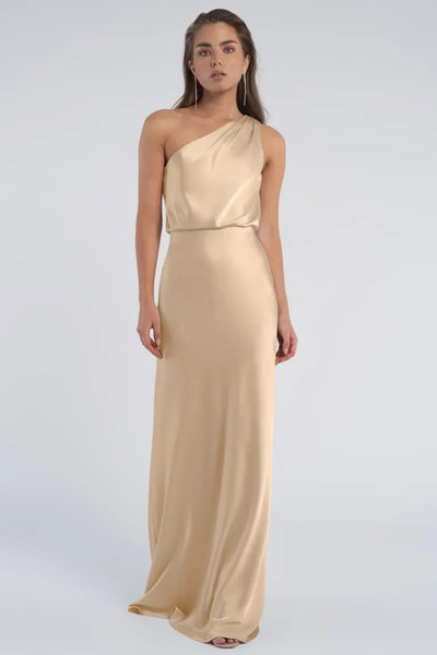 A woman in a beige one-shoulder Jenny Yoo Bridesmaid dress crafted from luxe satin fabric stands against a plain background.