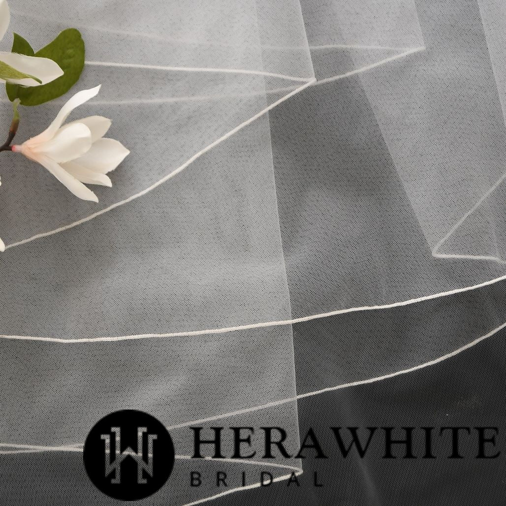Close-up of delicate ivory single tiered bridal veil fabric with sheer layers, accompanied by a small branch with white flowers. The soft tulle veil features serged edging for an elegant finish. The text "Bergamot Bridal" is displayed in the lower right corner.