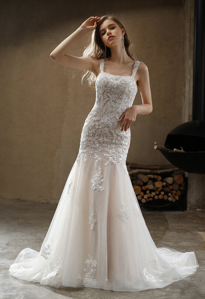 A woman in an elegant Square Neckline with Lace Straps Mermaid Wedding Gown by Bergamot Bridal, posing in a room with rustic decor.