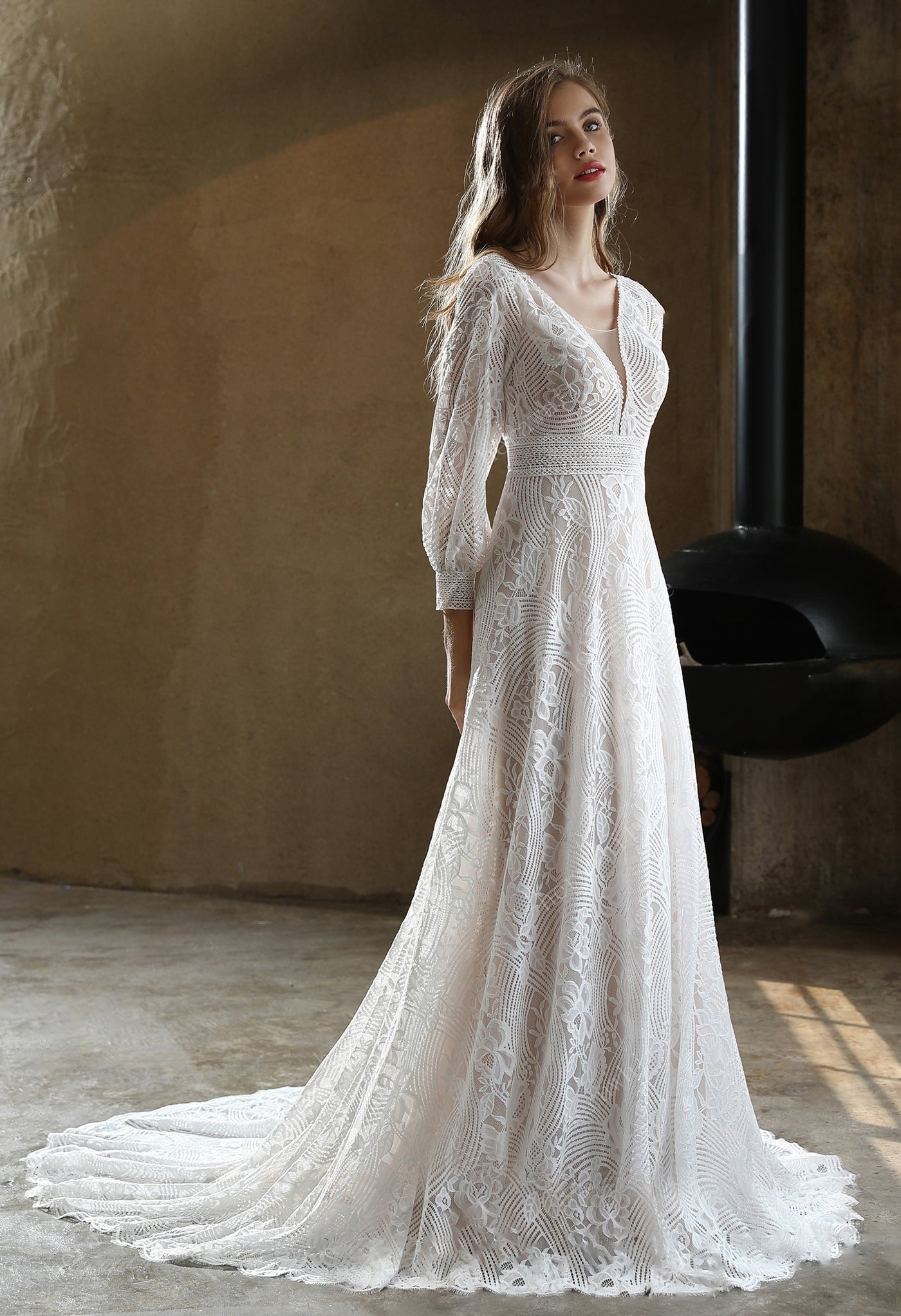 A woman in a white Bergamot Bridal Plunging V-neck Lace Long Sleeve Bohemian Wedding Gown is standing in a room.