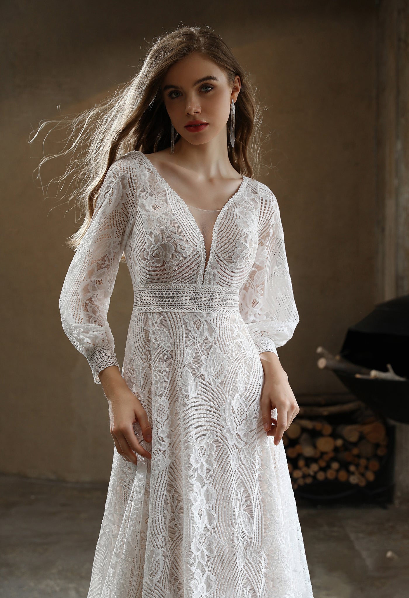 A woman in a Plunging V-neck Lace Long Sleeve Bohemian Wedding Gown by Bergamot Bridal is standing in front of a fireplace.