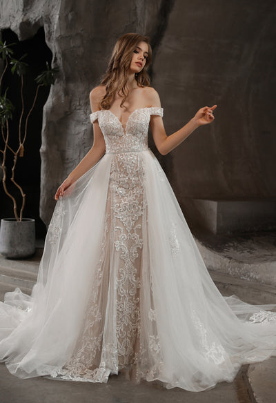 A woman in a Bergamot Bridal Gorgeous Lace Fit and Flare Bridal Gown with Detachable Train stands against a textured backdrop.