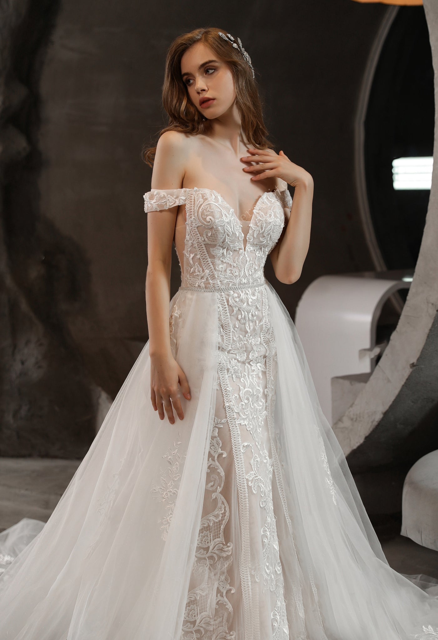 A woman in an elegant off-the-shoulder wedding dress with lace accents posing in an indoor setting. 
Product Name: Gorgeous Lace Fit and Flare Bridal Gown with Detachable Train by Bergamot Bridal.