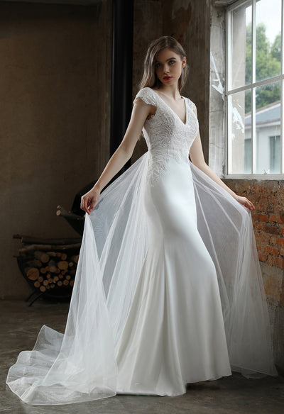 A woman in an elegant white Bergamot Bridal crepe sheath wedding dress with lace cap sleeves and detachable train poses in an industrial-chic setting.