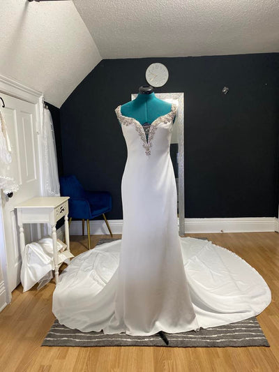 A white Pronovias Newton Gown - Off The Rack fit and flare dress with elaborate beading on the neckline displayed on a mannequin in a room with dark walls and a blue chair.