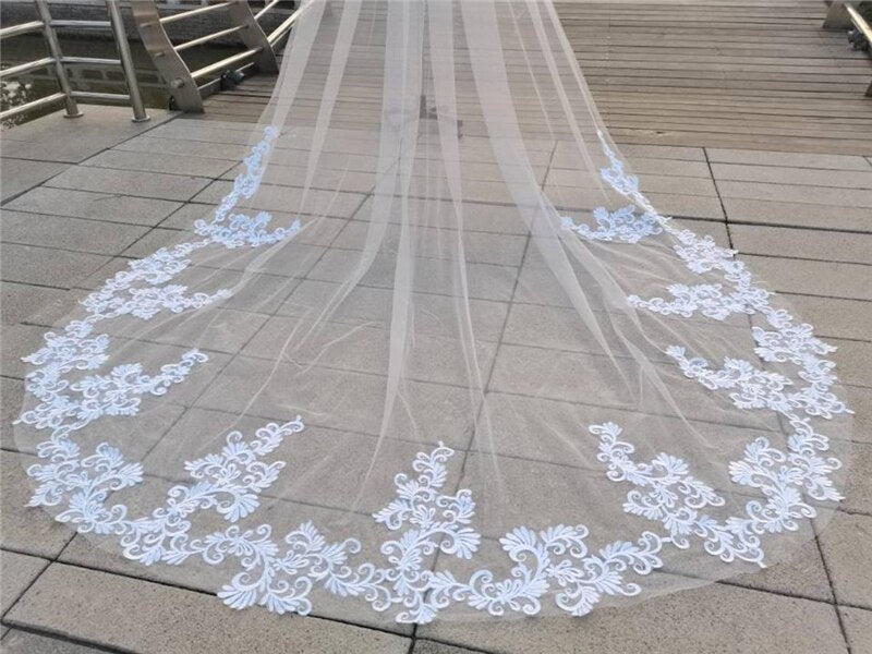 A long bridal veil with delicate white lace detailing from Bergamot Bridal's Lace Appliqued Cathedral Veil spread out on a wooden walkway.