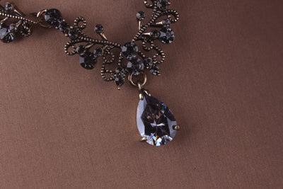 A Bergamot Bridal Baroque style black crystal bridal jewelry set necklace, earrings, and tiara pendant necklace with a large teardrop gem on a brown backdrop.