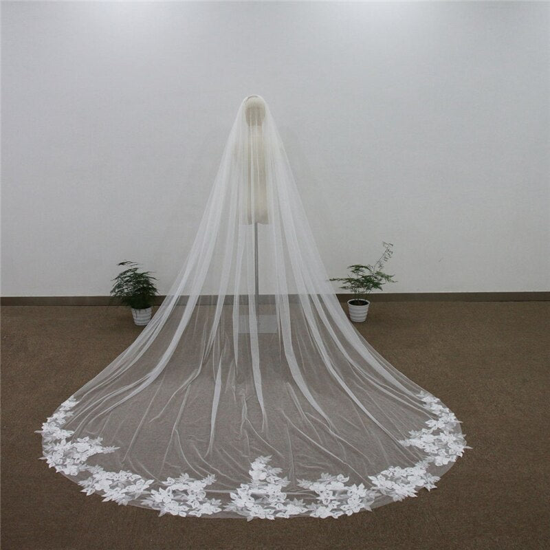 A bride in a long white gown and flowing veil stands with her back to the viewer in a simple room with two potted plants, showcasing one of the elegant wedding dresses from Bergamot Bridal featuring the Embroidered Floral Lace Cathedral Wedding Veil.