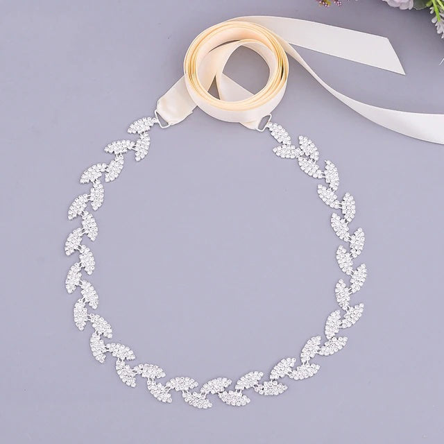Elegant bridal headband with leaf pattern made of sparkling crystals, displayed on a light grey background with ivory ribbon tied at the ends.