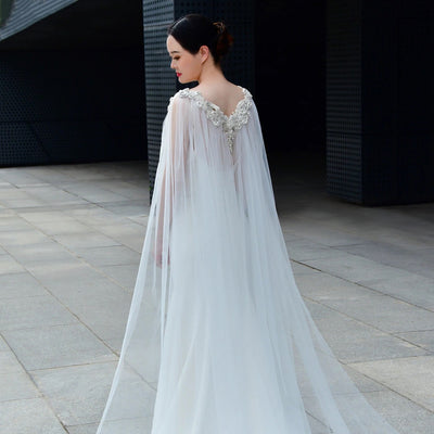 A woman in an elegant white gown with ornate neckline and a Bergamot Bridal Bridal Cathedral Length Cape with Beaded Detail, standing outdoors on a gray pavement with a modern building background, perfect for showcasing at bridal shops in London, Ontario.